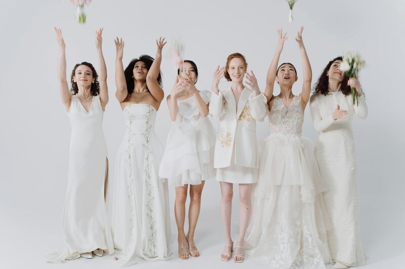 Six women in elegant white dresses joyfully tossing flower bouquets during a wedding celebration photoshoot