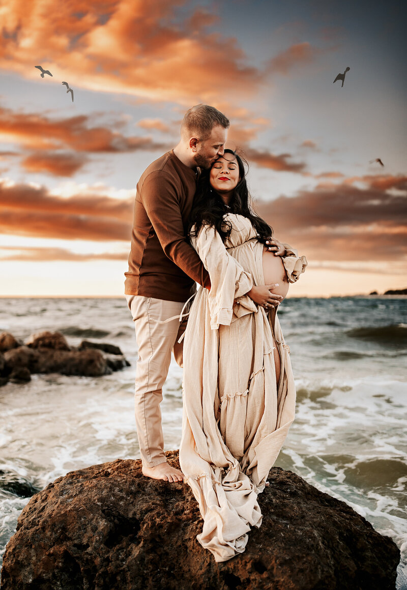 Maternity Photographer, a man and his pregnant partner embrace at the beach, they stand in shallow tides