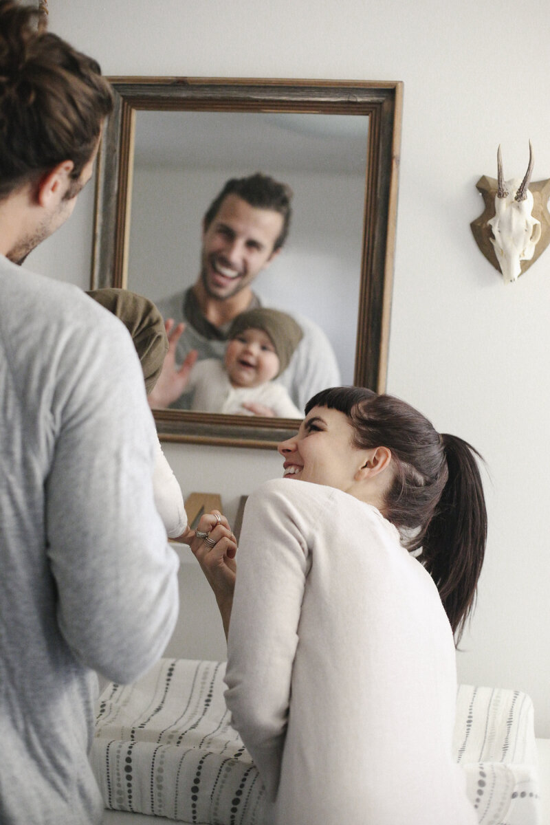 A woman and man stand beside a baby changing table, smiling and looking into a wall mirror that reflects their happy faces and the baby in a knit hat.