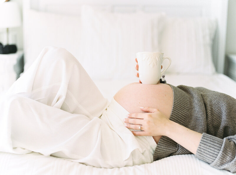 Pregnant woman's belly balancing a cup of tea while lying on a white bed.