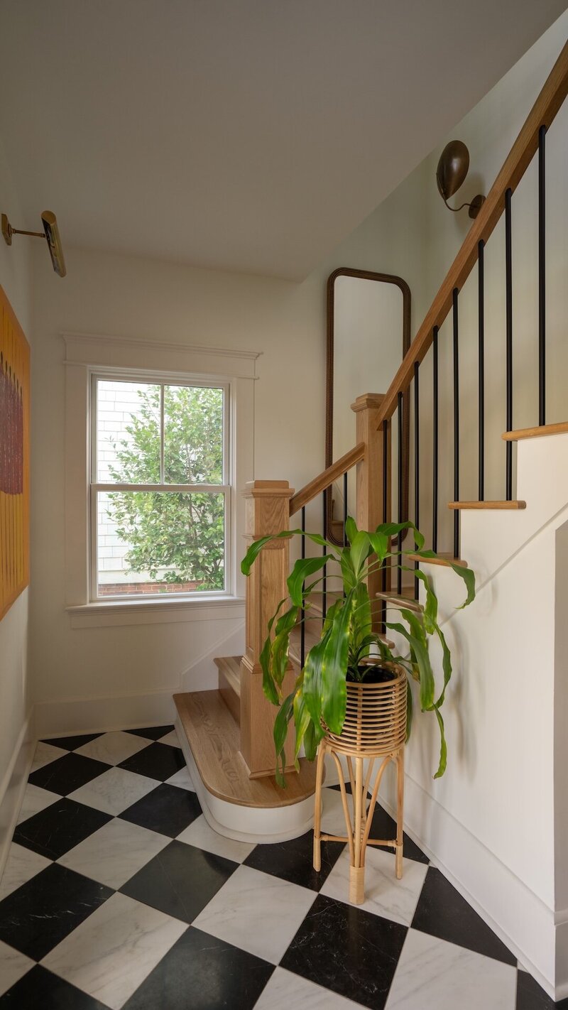 Entryway with black and white checkered tile floor, wooden stair bannister, and rattan plant pot adding warm natural tones.