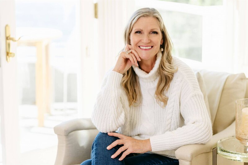 Phillipa Leseberg with long hair, dressed in a white sweater, sits in a chair smiling and resting her chin on her hand. The bright window behind suggests inspiration—perhaps she's lost in thought about her next book.
