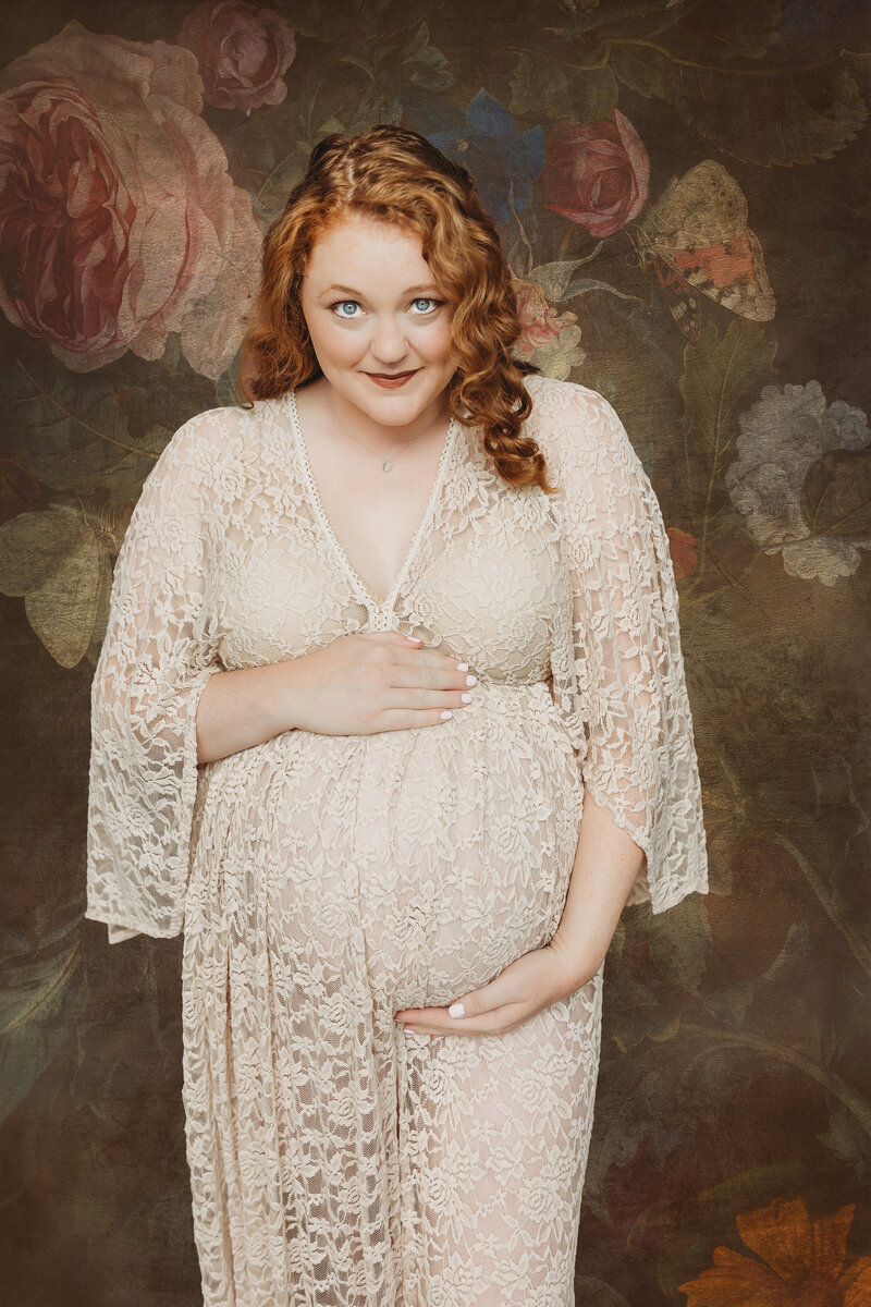A maternity photo depicting a pregnant woman wearing a dark velvet dress, standing outdoors. She is cradling her belly with one hand while her partner, dressed in a light jacket, places his hand on her belly, signifying affection and support.