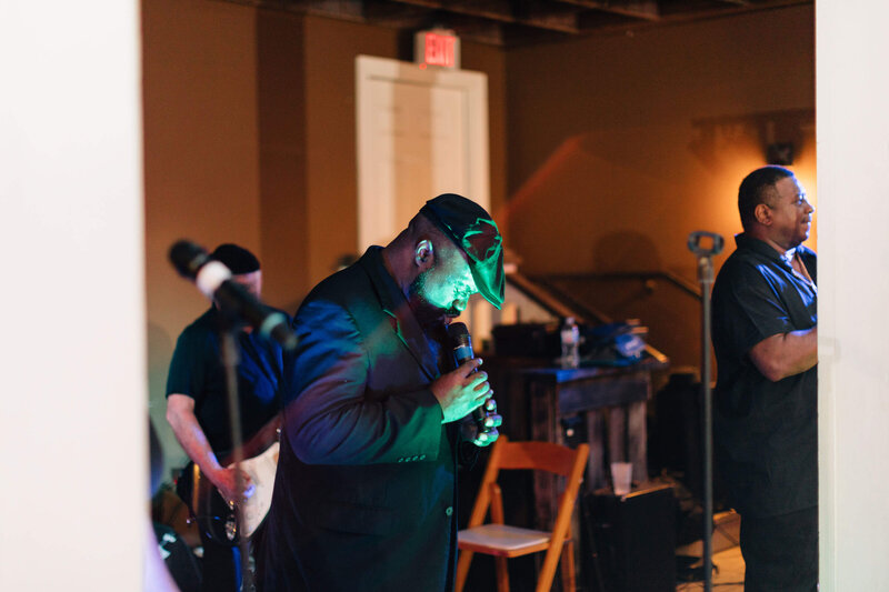 A band performs indoors; a man in a dark jacket and hat sings into a microphone at center and a musician plays guitar in the background under warm lighting.