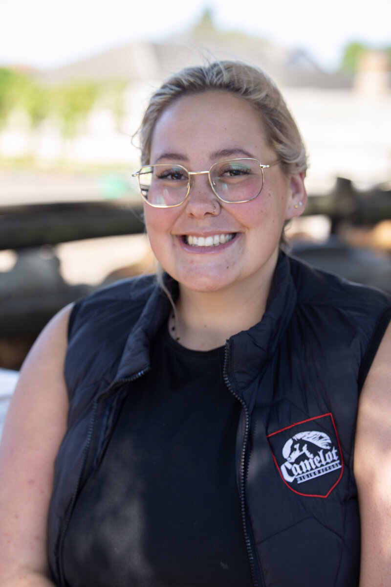 Robyn Theron, instructor at Camelot Horse Riding School, smiling while wearing a Camelot-branded vest beside a horse.