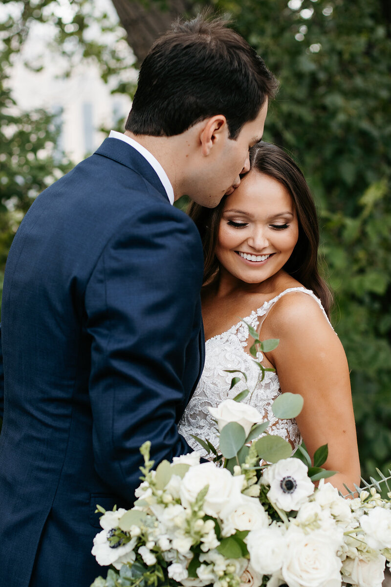 Groom kissing bride on the forehead while she looks down at her bouquet of white and green flowers