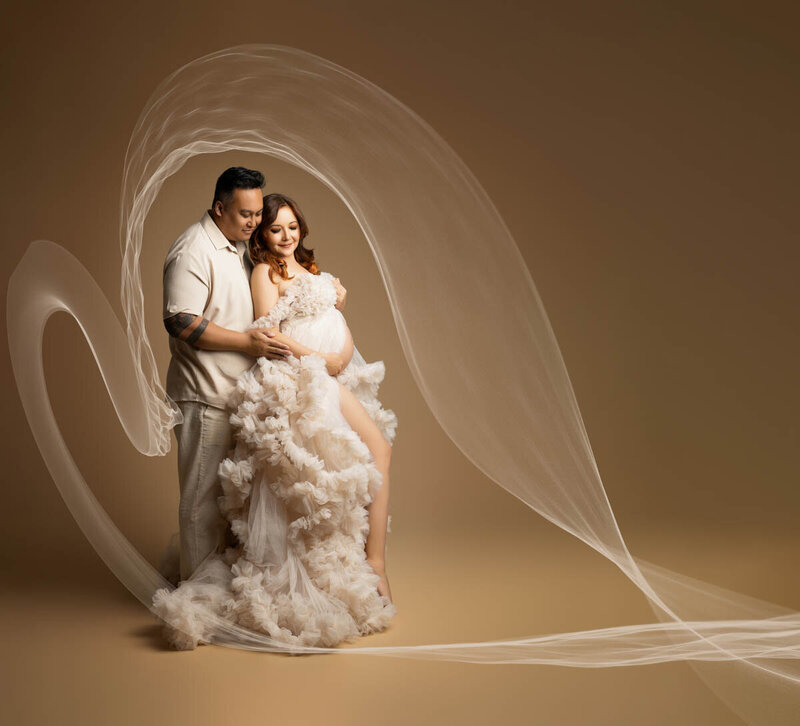 Couples maternity portrait of a woman dressed in a white tulle gown and a man dressed in beige both posed in front of a brown backdrop