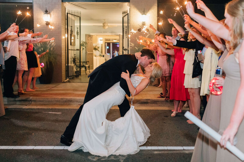 A bride and groom share a romantic kiss as the groom dips the bride on a street, surrounded by cheering guests holding sparklers during a nighttime wedding celebration.