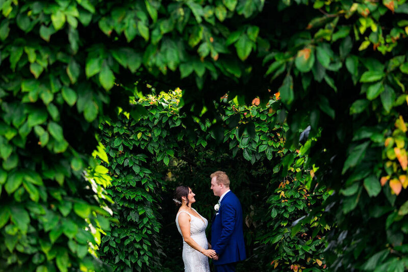A couple stands in very green leaf tunnel while looking at each other and holding hands