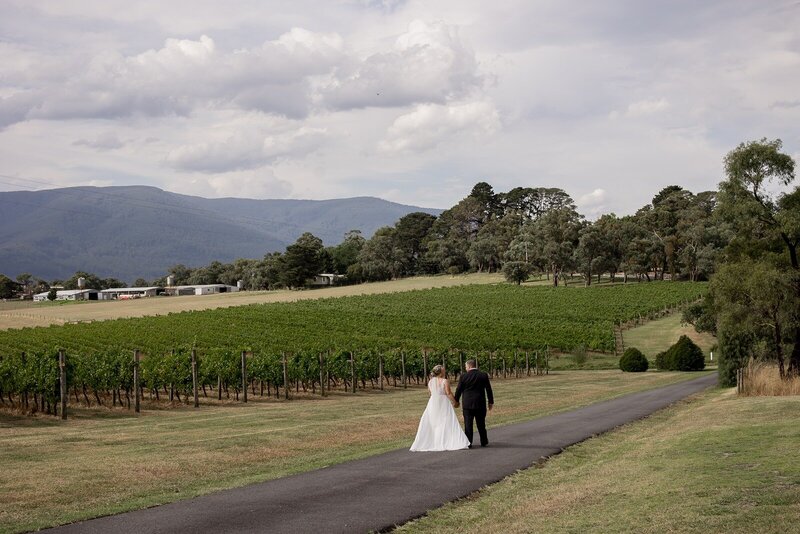 Yarra valley mountain scenery with vineyards, and a bride and groom walking in the foreground.