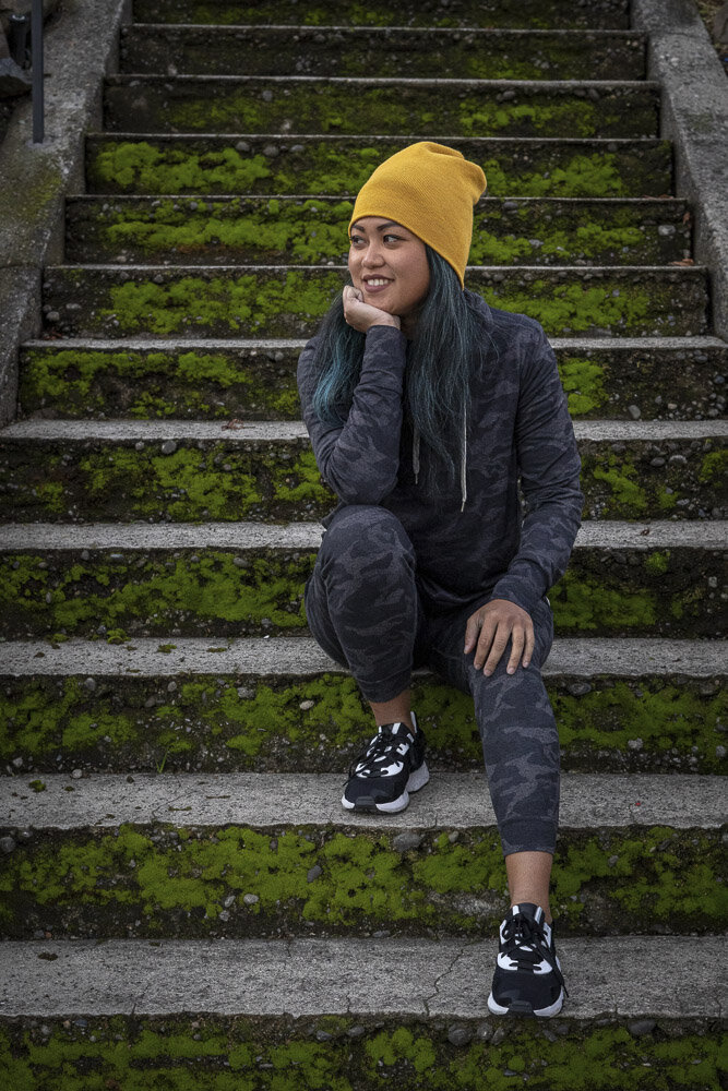 This image shows an Asian woman with long blue hair wearing a yellow beanie hat sitting on moss-covered steps. She rests her head in one hand, looking past the viewer, with a wide smile.