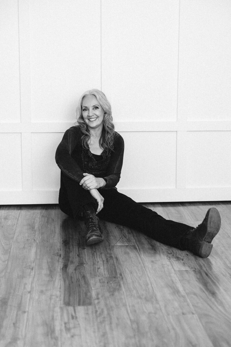 Black and white photo of a San Francisco Bay Area brand photographer  sitting against a white wall in her studio.