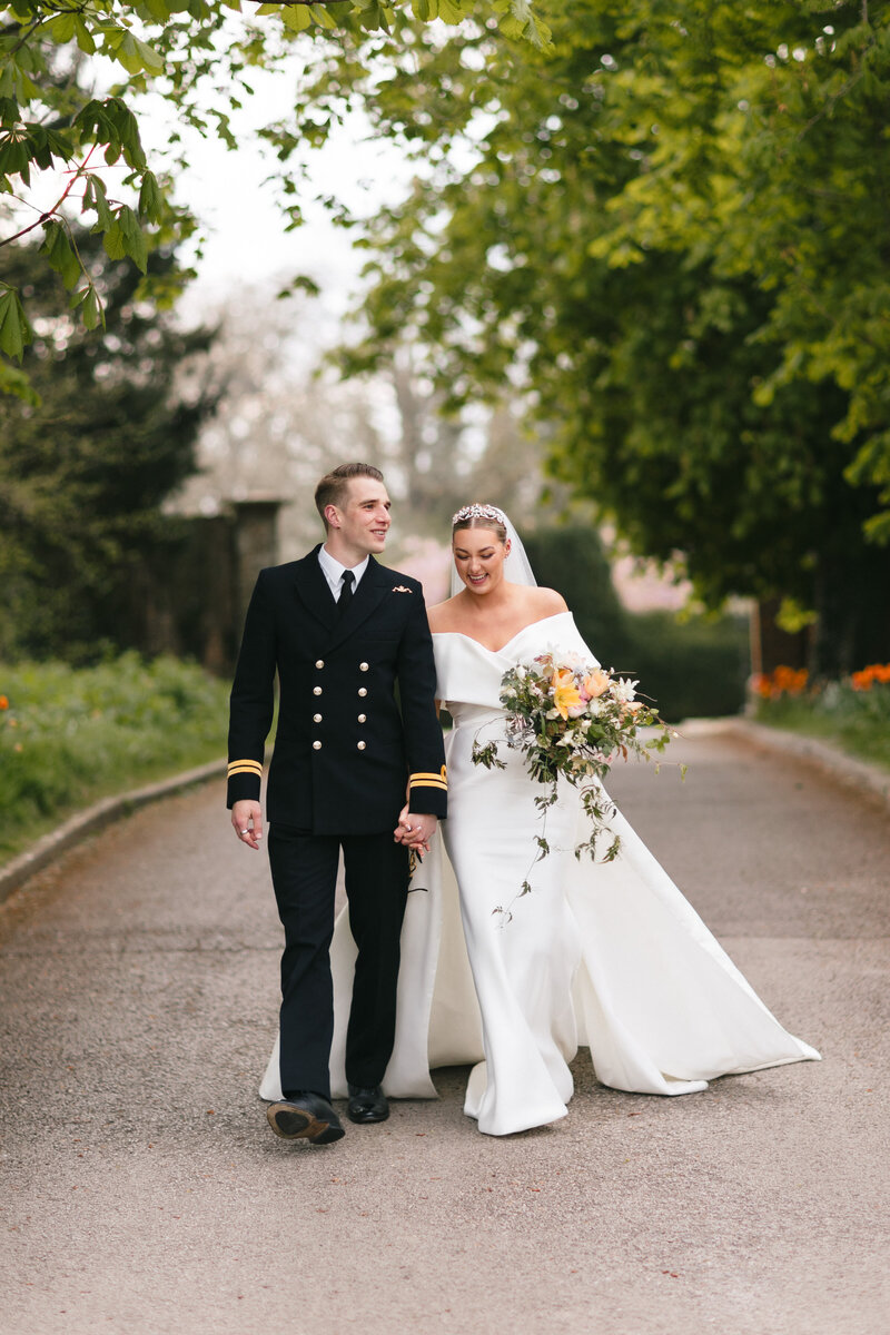 A bride and groom walk hand in hand on a tree-lined path. The groom is in a naval uniform, and the bride wears an off-shoulder white gown while holding a floral bouquet. Both are smiling amidst a lush green backdrop at Lainston house
