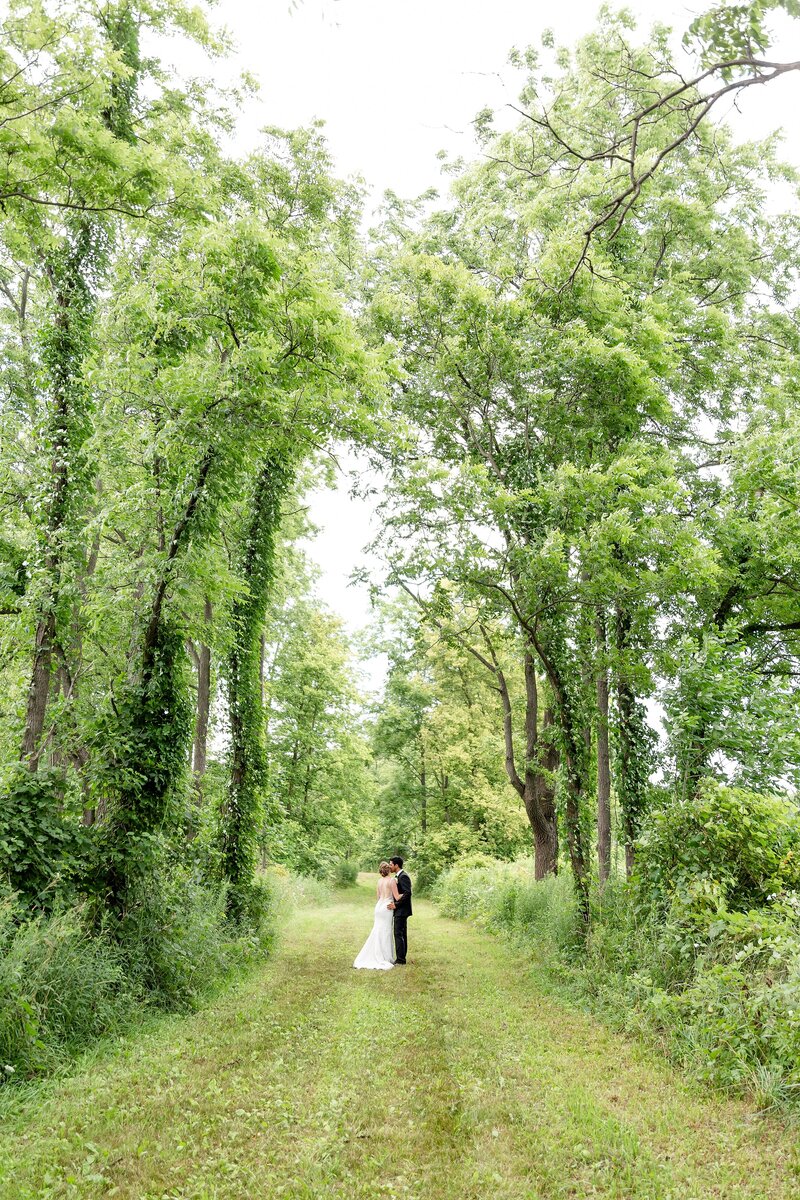 Newlyweds embrace under a large archway of trees at her grandmothers farm during their wedding in mount brydges