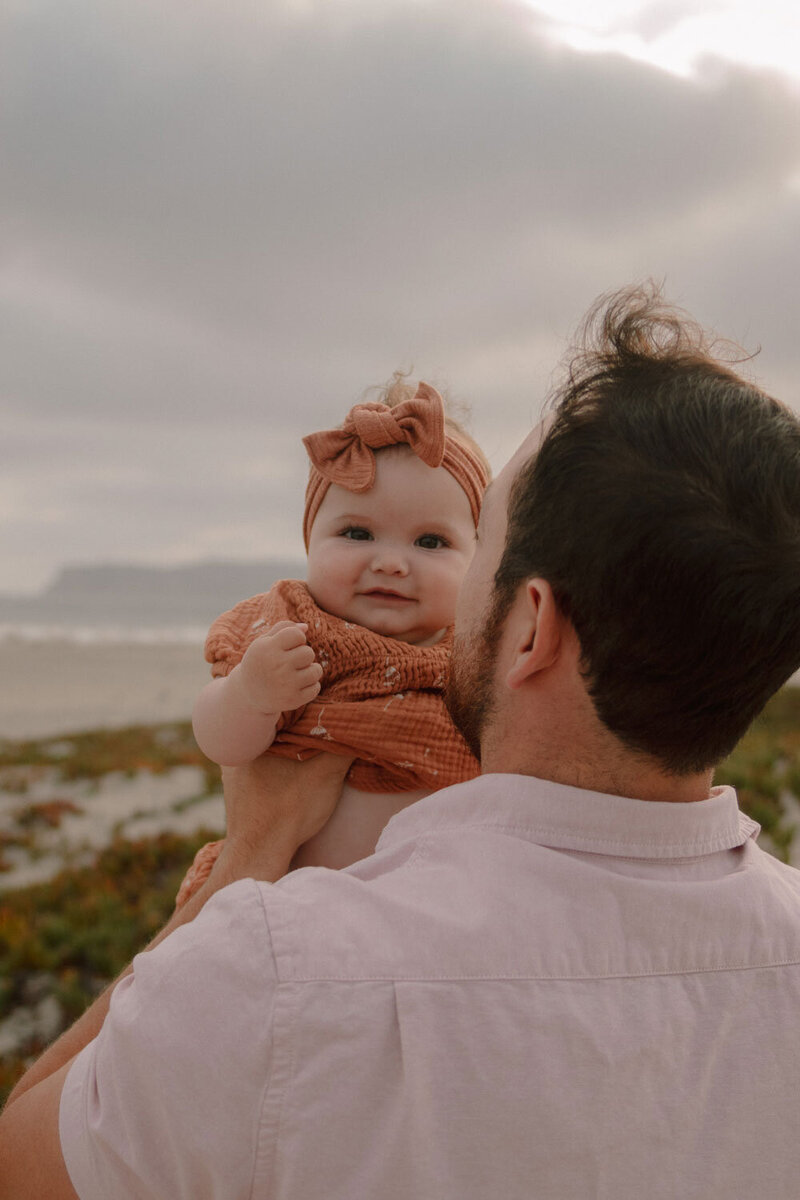 Baby girl looking over her father's shoulder at the beach, with a soft sky in the background
