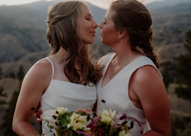 2 brides slowly embracing for a kiss on a mountain scape holding a bouquet of flowers.