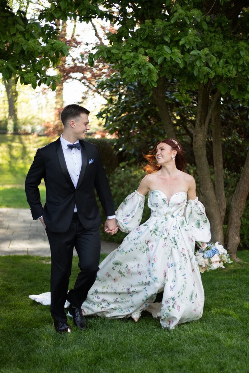 Elopement Wedding Packages Rhode Island | Kelsey Sheehan Photography Timeless Rhode Island Weddings | A joyful couple walks hand in hand on grass, the groom in a black tuxedo, and the bride in a floral gown, with trees and sunlight in the background.
