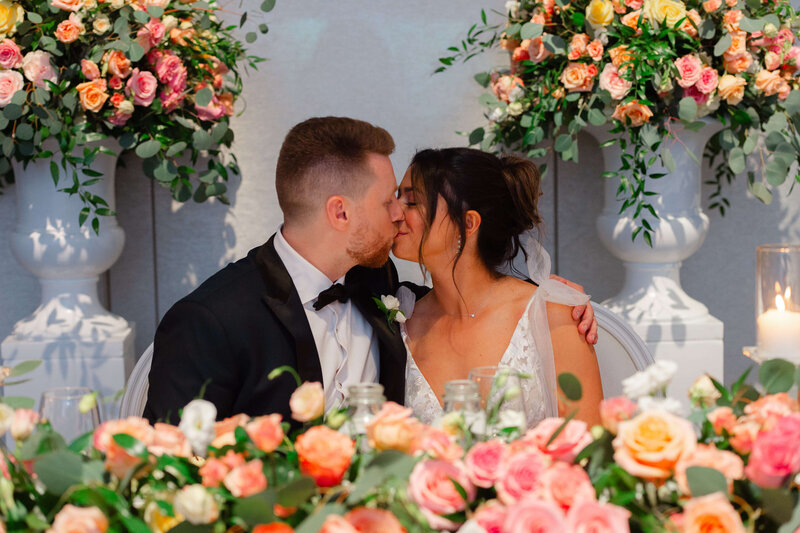 bride and groom kiss at their head table surrounded by florals during their Chateau Le Parc wedding reception.  Taken by Ottawa wedding photographer JEMMAN Photography