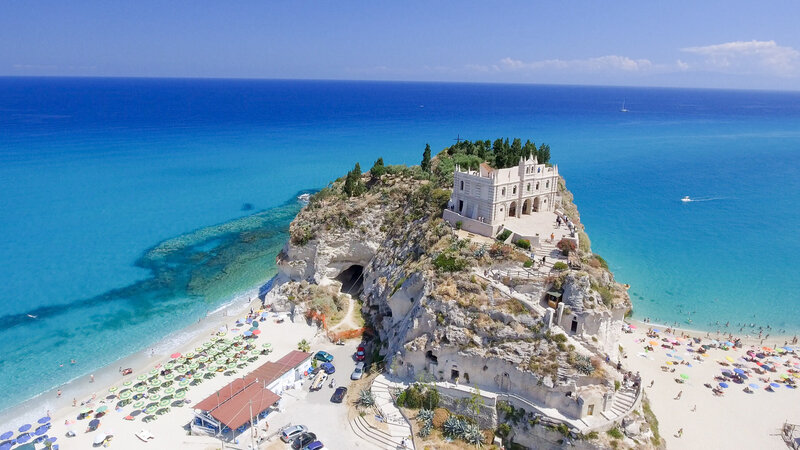 Aerial view of a seaside cliff with a historic building, reminiscent of luxury vacations, surrounded by turquoise waters and sandy beaches filled with colorful umbrellas and sunbathers. Lush greenery tops the cliff under a bright blue sky.