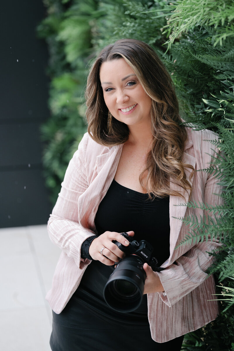 Stephanie looking at the camera smiling posed with her camera and dressed in her signature pink color top