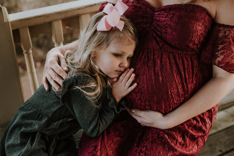 Maternity photo of young daughter in gray dress with pink bow laying on pregnant moms belly while mom is sitting on bench in red dress holding her pregnant belly and hugging daughter in Annapolis Maryland.