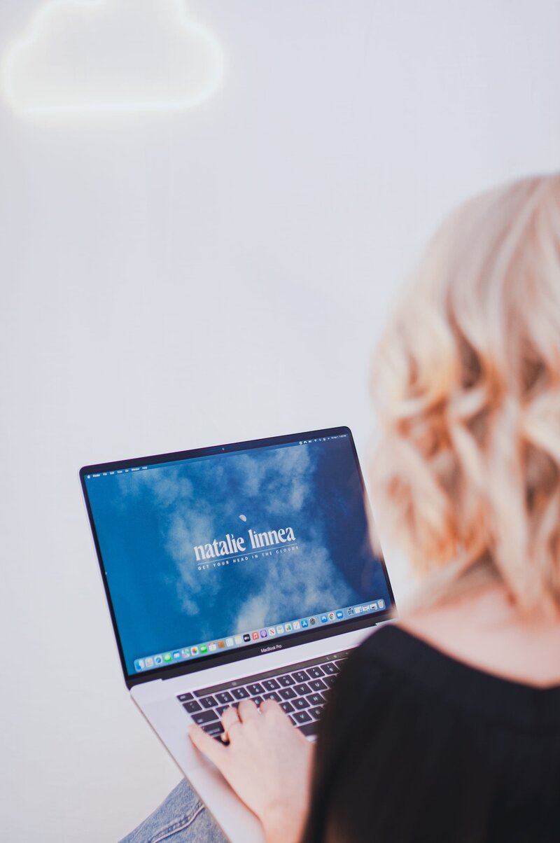 Back of woman's head and a laptop screen with an image of a sky and text that reads "Natalie Linnea"