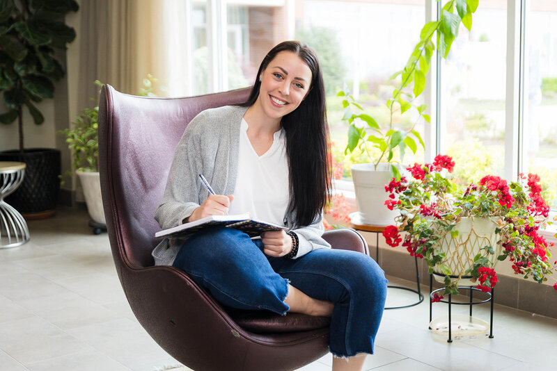 Woman sitting on the couch necxt to a furry blanket and plant with a stack of books in her lap smiling