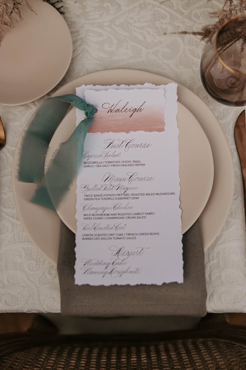 Hand-lettered wedding menu tied with a green ribbon, styled on a taupe plate with neutral-toned flatlay decor.
