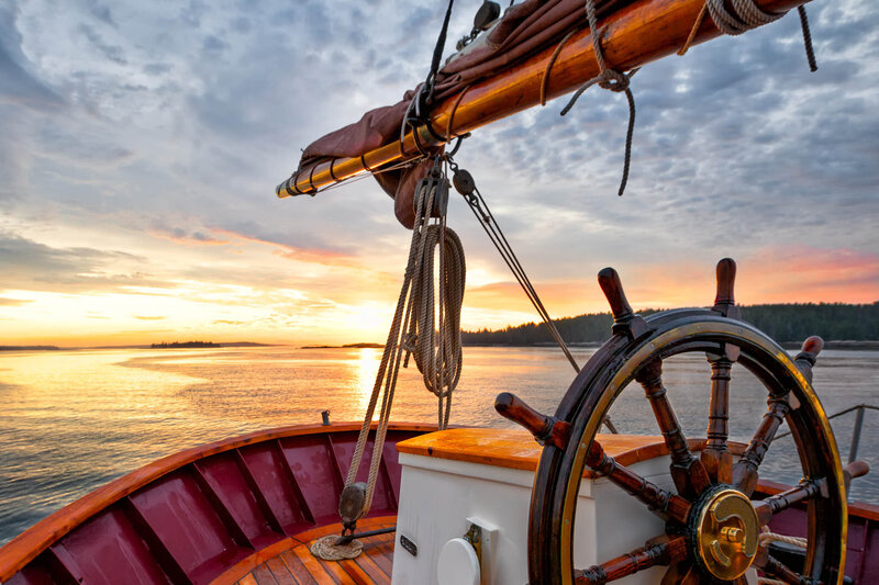 Wooden sailboat helm and rigging at sunset, overlooking calm waters and distant shoreline under a colorful sky.
