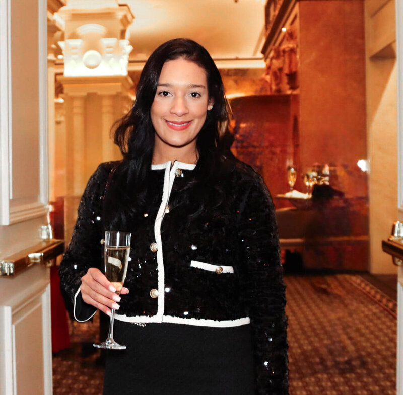 Woman with long dark hair wearing a black sequined jacket with white trim, holding a glass of champagne and smiling in an elegant indoor setting.