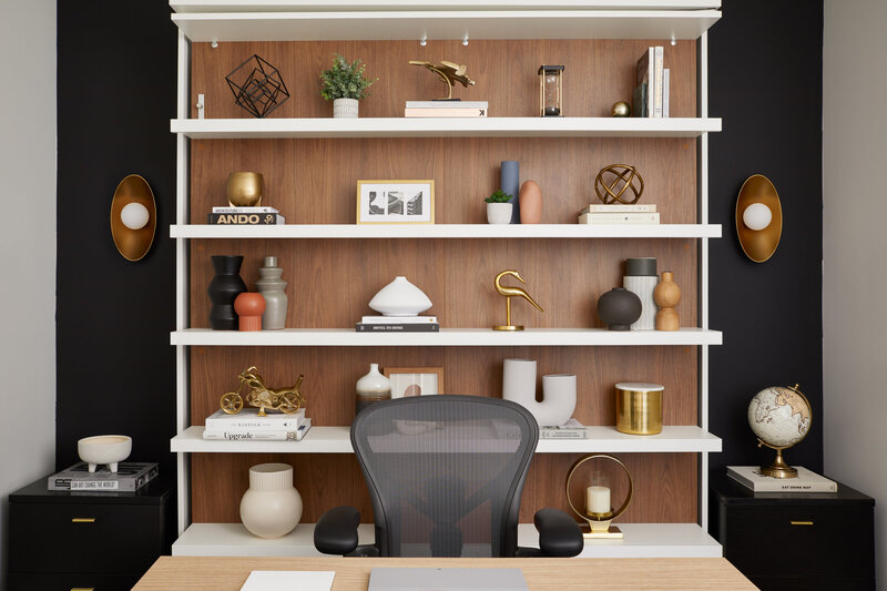 A modern, stylish office space featuring contrasting black, white tones and wood accents. The office contains a desk, a mesh-back chair, and elegant white and wood shelves showcasing various art pieces.