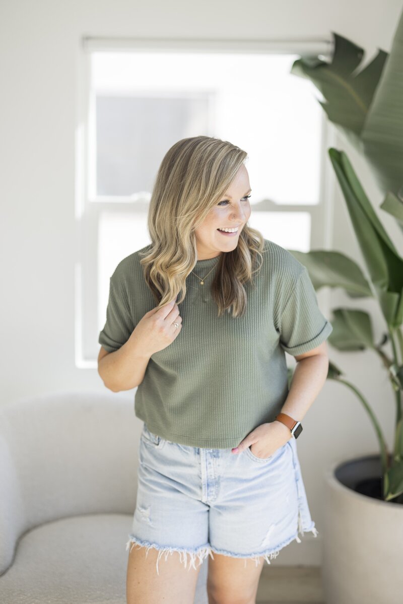 Krista Marie, a brand photographer for women, standing in front of a window and green plant smiling and looking away from the camera