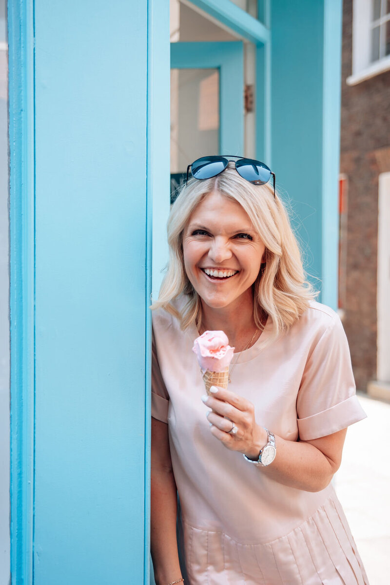 Emily Collins, mindset coach, smiling while holding a pink ice cream cone and standing near a bright blue doorway.