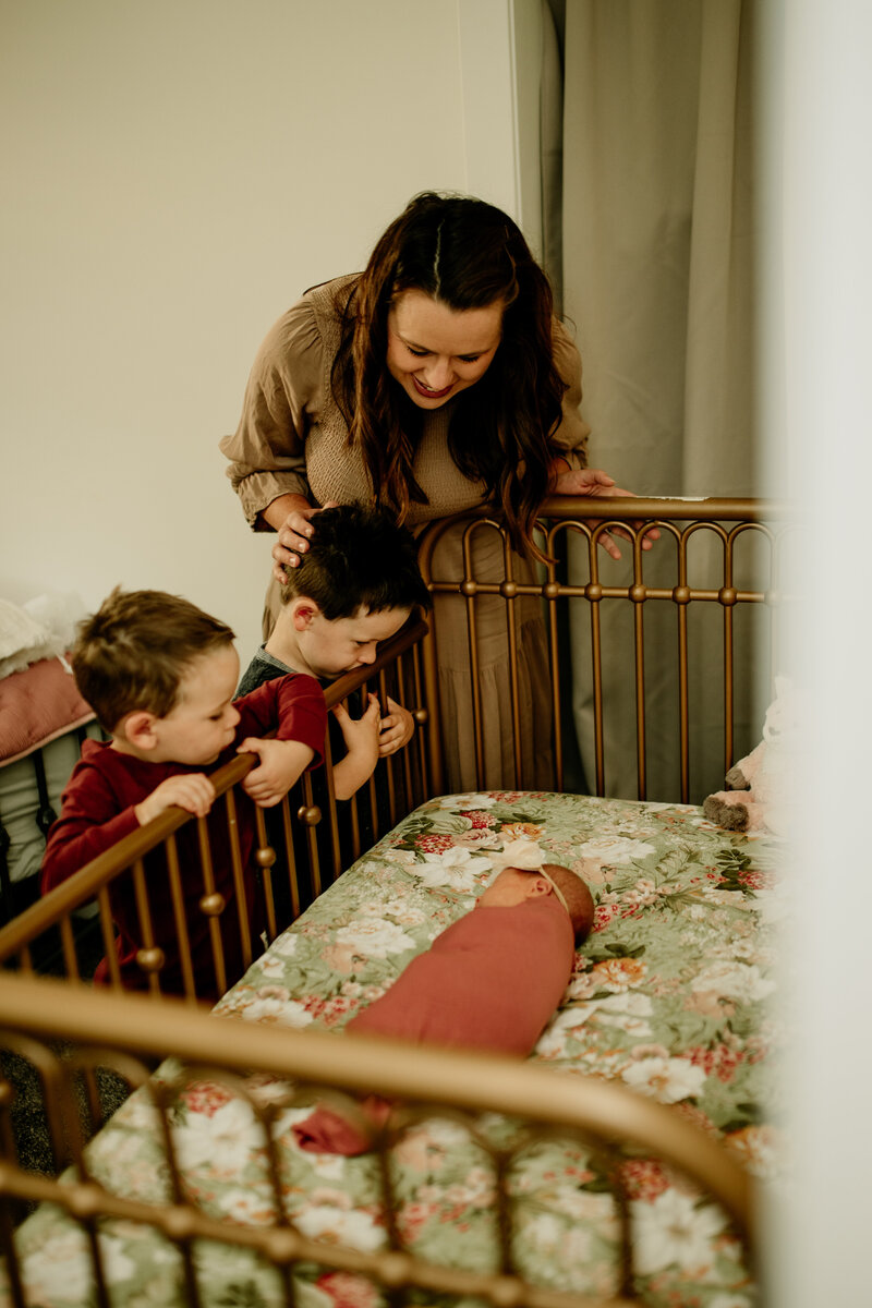A mom and her two toddlers peek into a crib where their baby sister rests peacefully during an in-home newborn family session. 