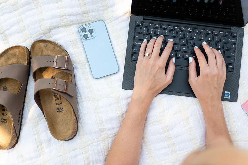 A virtual assistant types on her computer with her phone beside her, laying on a picnic blanket