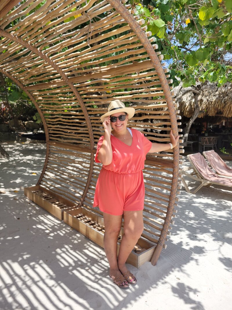 Woman in hat, sunglasses and orange outfit under bamboo arch on shaded beach in Curacao