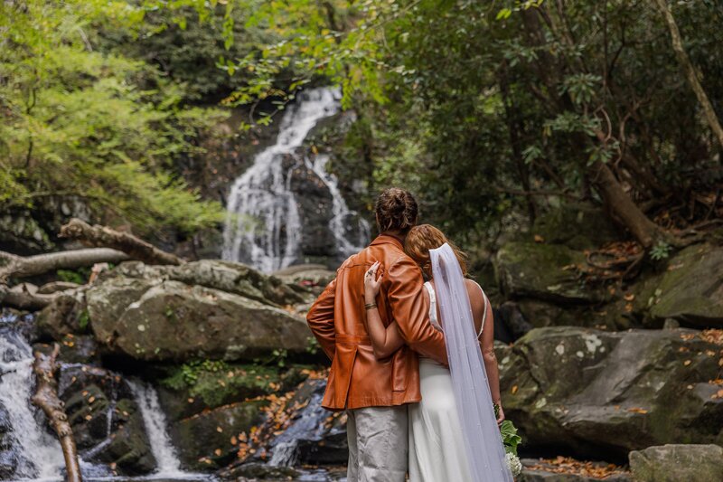 A bride and groom stand in front of a large, tumbling waterfall with their backs facing the camera. The are looking out at the waterfall with their arms around each other. The bride is in a flowy wedding dress with a long veil and the groom is wearing tan pants with an orange leather jacket. They are surrounded by rocks and lush green trees.