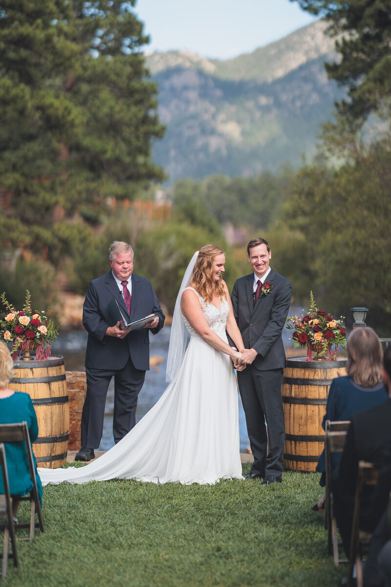 A bride and groom holding hands during their wedding ceremony next to a river in the woods in Estes Park
