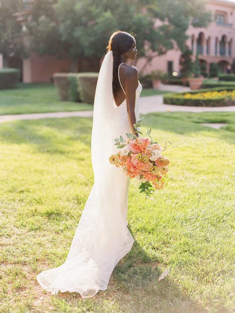 A bride stands on a green lawn, the the sun shining on her. She has her back facing the camera showing off her low back, body fitting gown, with a short train, and long veil. At her side she holds a large fluffy pink and coral bouquet.