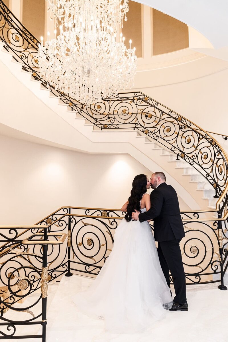 A bride in a flowing white wedding dress and a groom in a sleek black tuxedo share an intimate kiss on a sweeping staircase with ornate black and gold railings. The glow of an opulent crystal chandelier casts a warm light over the scene, highlighting the elegance of their wedding day