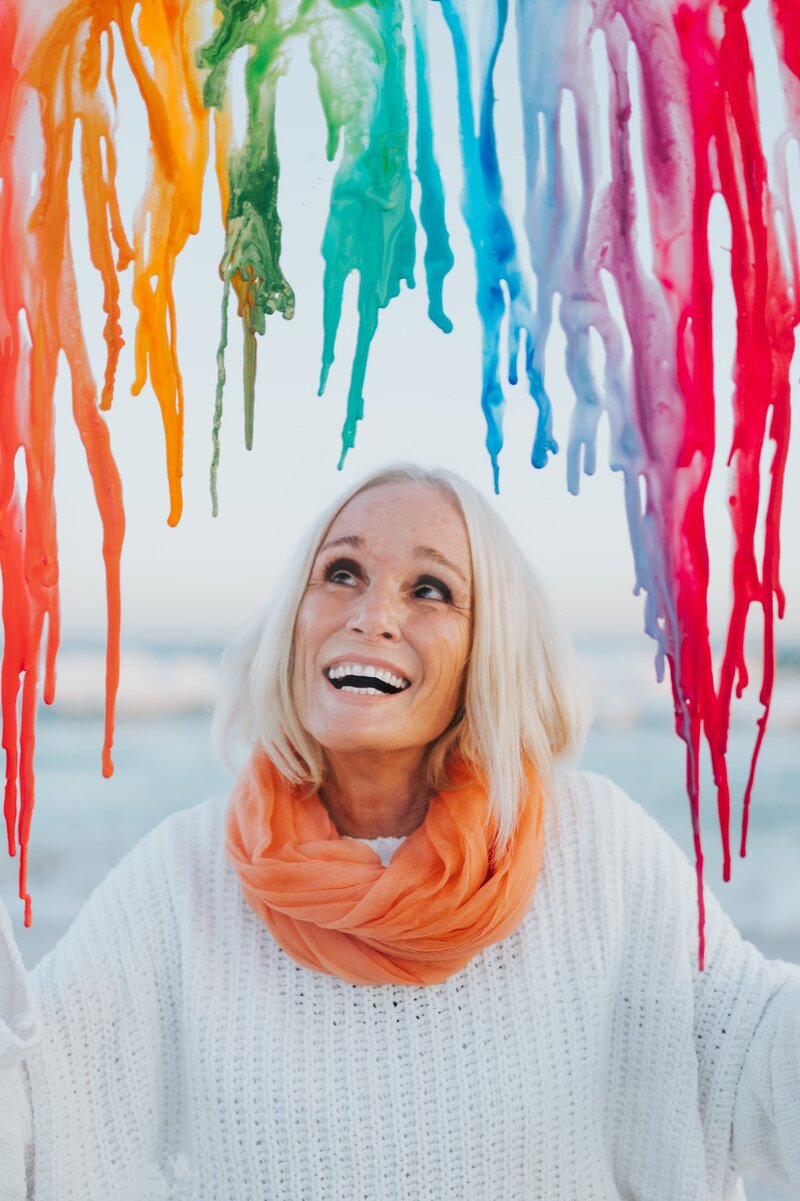 Older woman with white hair wearing a white long-sleeve shirt and orange scarf smiles up at melted rainbow crayons.
