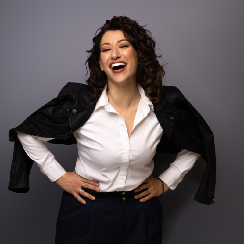 Woman in a white shirt and black jacket with her hands on her hips laughing
