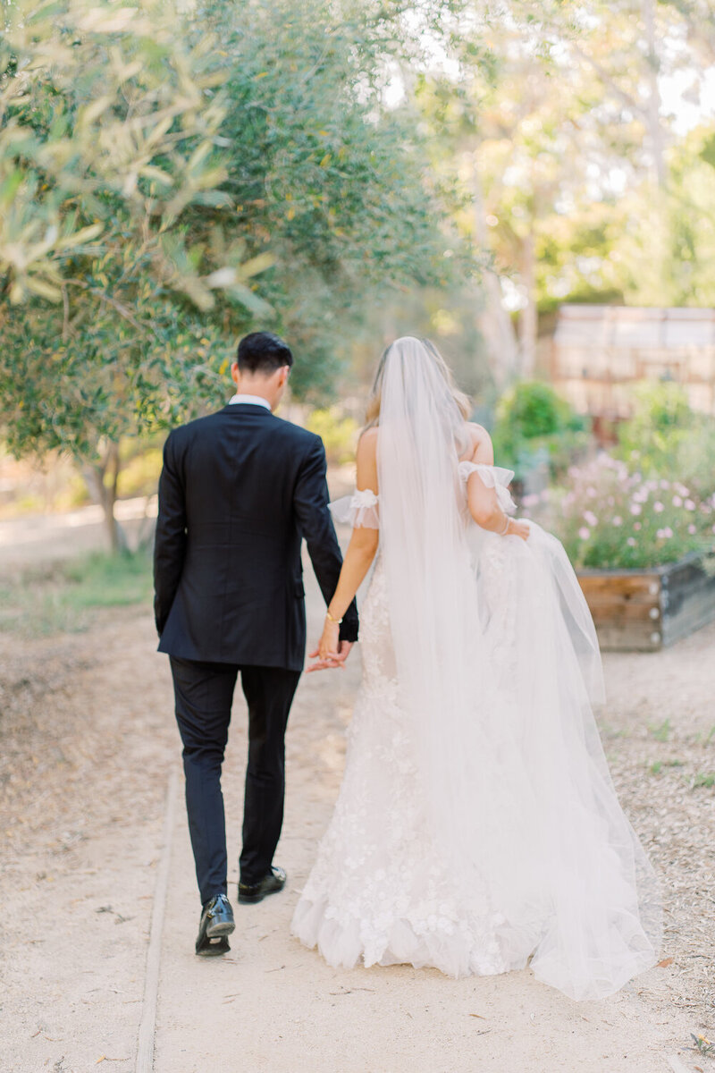A bride and groom walk hand with there backs to the camera. The groom's suit is all black, and the bride has a long toile veil.