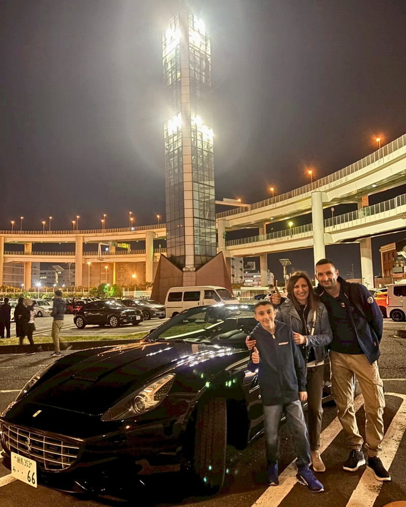 A family of three smiles beside a sleek black Ferrari at the Daikoku Parking Area in Japan, surrounded by city lights and expressway overpasses at night.