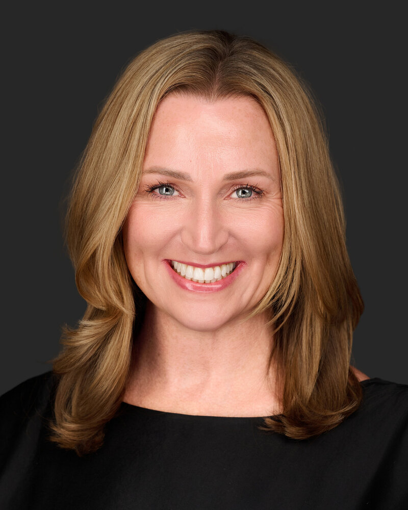 Jenn Cloutier, self-leadership coach, smiling warmly with shoulder-length blonde hair, wearing a black professional top against a dark gray background.