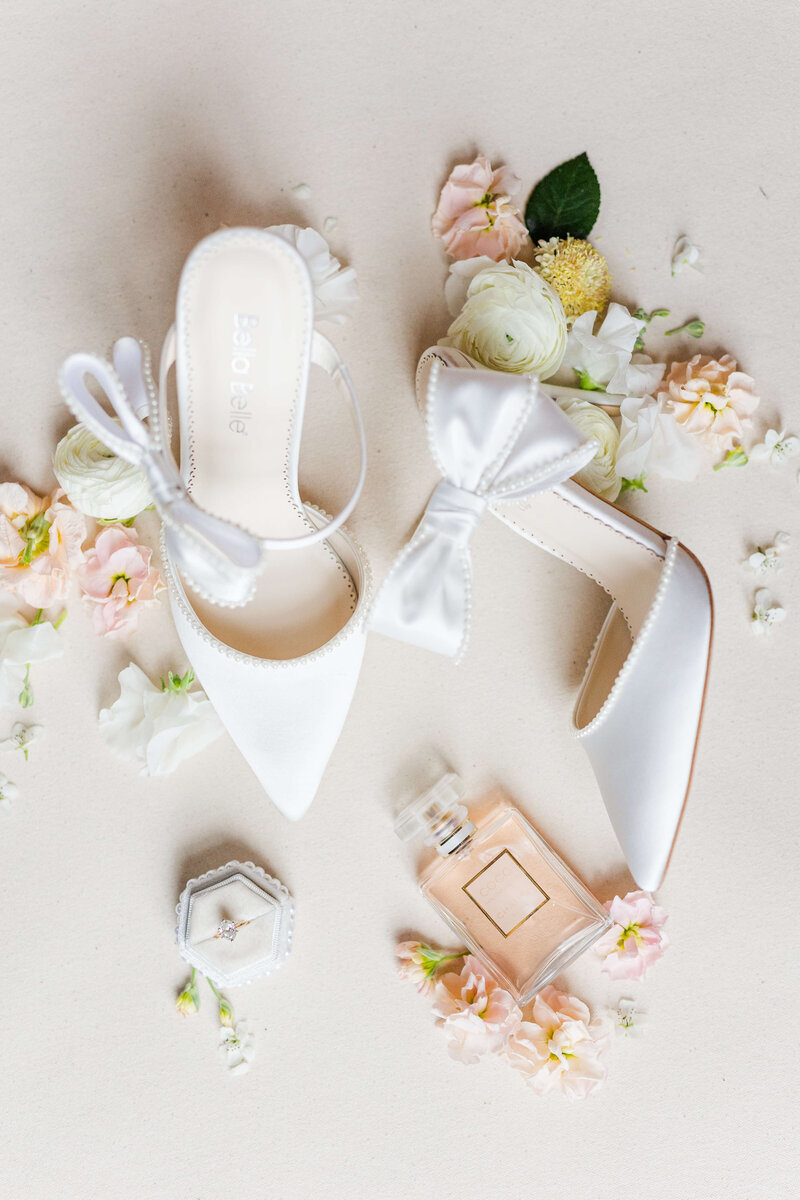 An up close photo of a bride's shoes. They are white pointed heels with a bow. There are spring flowers around them along with an engagement ring and a bottle of pink perfume.