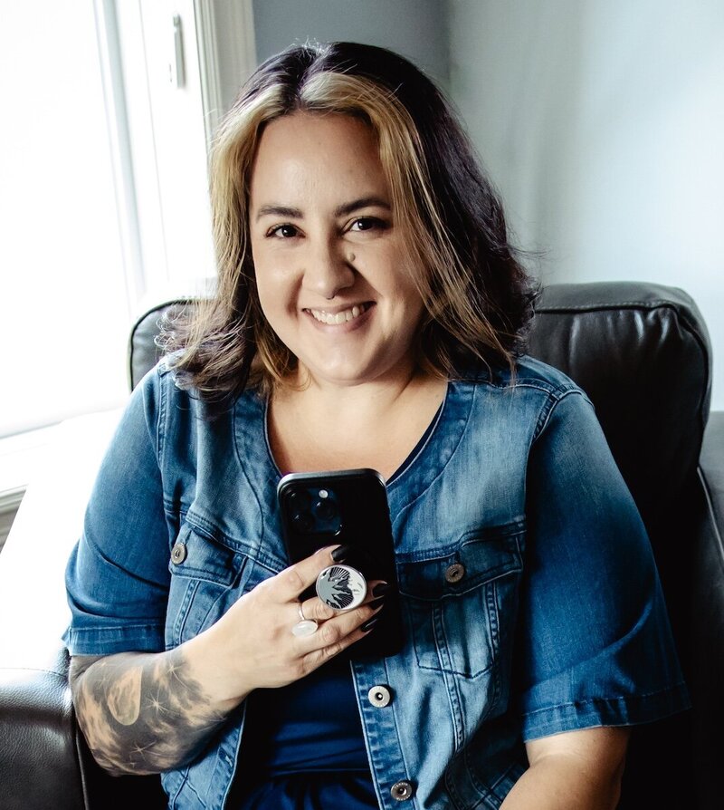 woman smiles in a jean jacket on a chair with her cell phone in her hand 