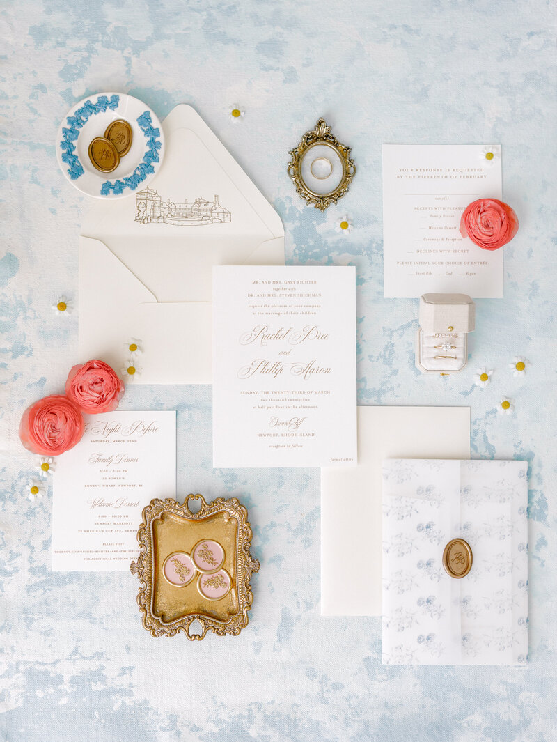 Flatlay of ivory wedding invitation with coral flowers, gold accents, and a drawing of OceanCliff Hotel on the inside envelope.