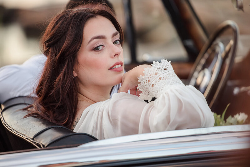 Bride gazes back from a vintage convertible, her lace cuffed sleeve resting elegantly as soft sunset light illuminates her features.