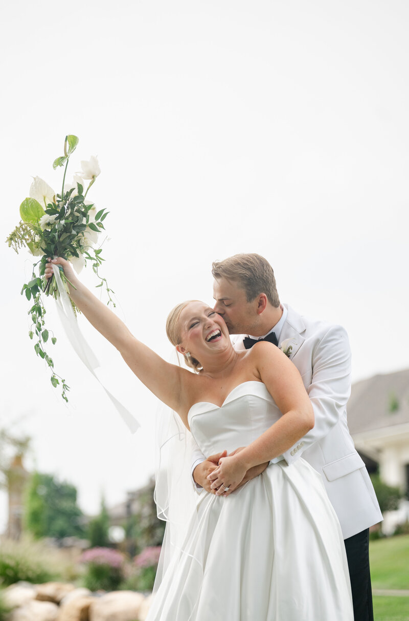 A bride cheers with her flowers in the air while her groom kisses her